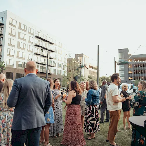 A crowd mingles at an outdoor event in a courtyard, with modern buildings, string lights, and lively summer conversation.