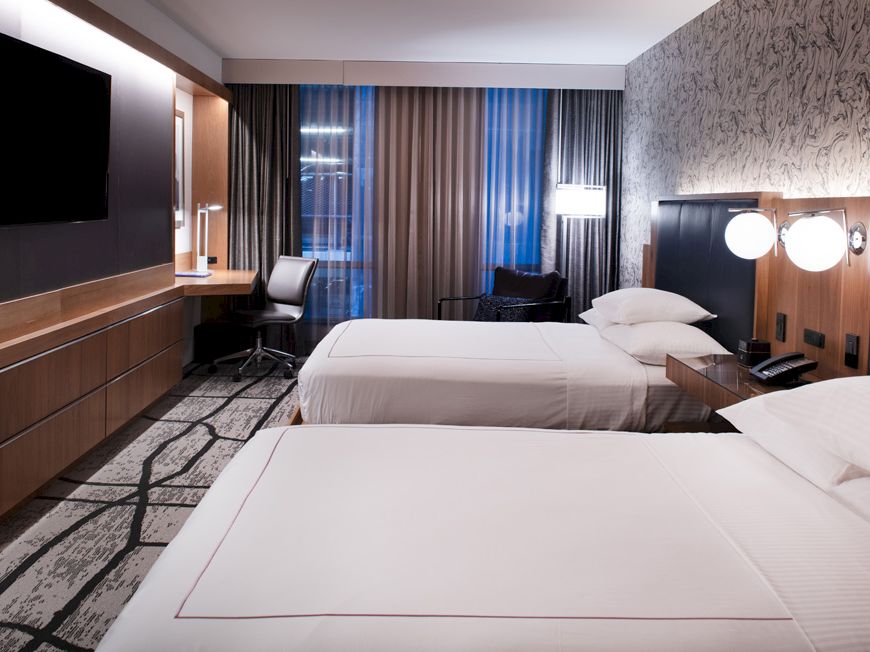 The image shows a modern hotel room with two beds, a flat-screen TV, a desk, and a window with curtains, all in a sleek, contemporary style.