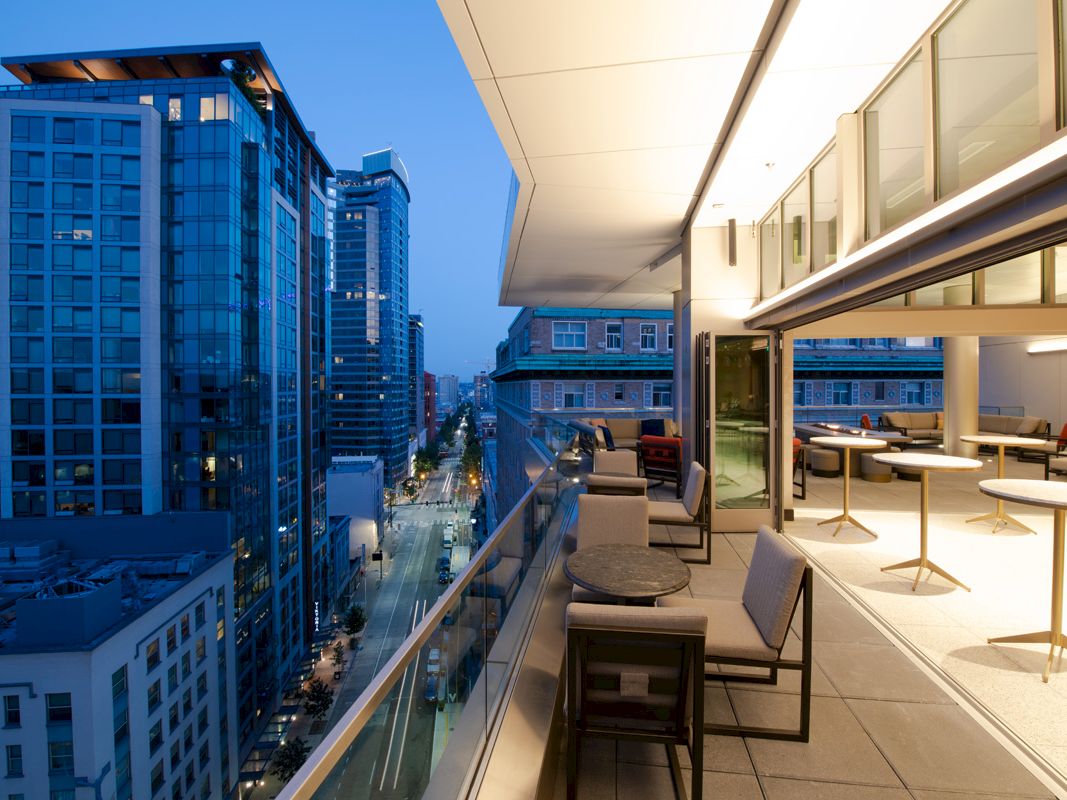 A modern cityscape at dusk with a view from a balcony showcasing tall buildings and a stylish outdoor seating area with tables and chairs.