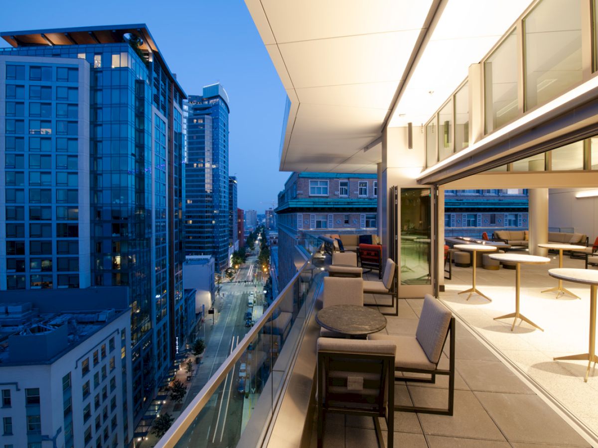 A modern rooftop terrace with seating overlooks a city street lined with tall buildings at dusk, creating a stylish urban atmosphere.