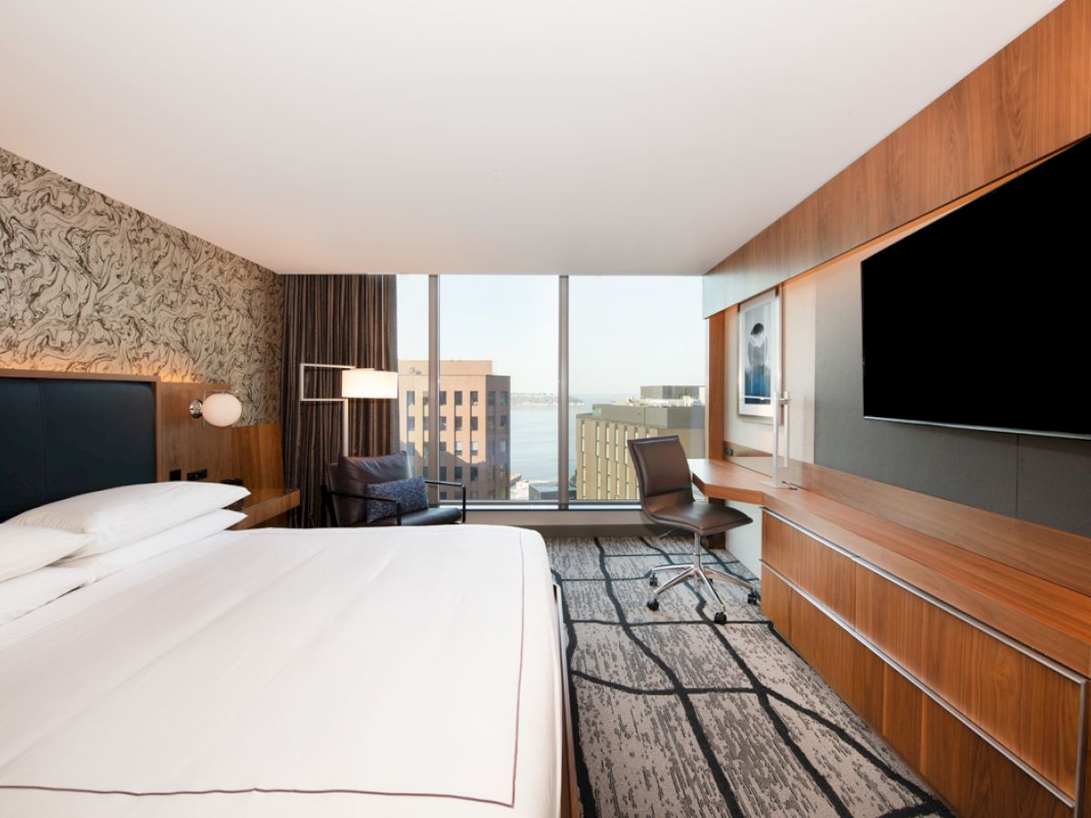 A modern hotel room with a large bed, wall-mounted TV, desk, chair, and window offering a city view.