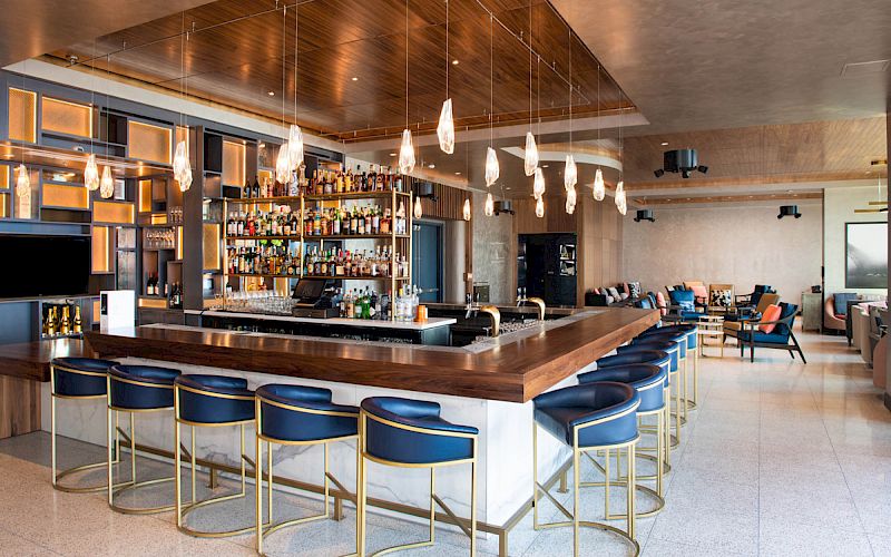 The image shows a modern bar with a wooden counter, blue cushioned stools, overhead pendant lights, and shelves stocked with bottles.