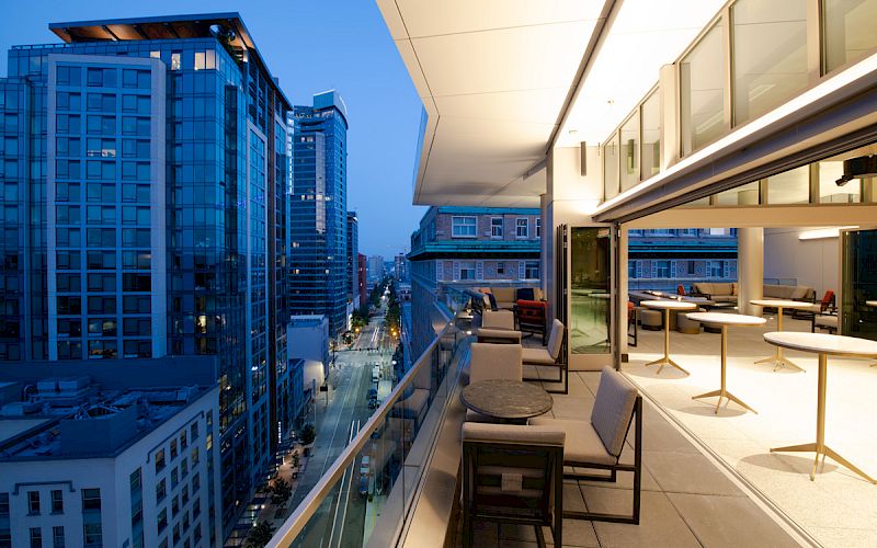 A modern balcony overlooking a cityscape at dusk, featuring seating areas and tables, with tall buildings and streetlights in the view.